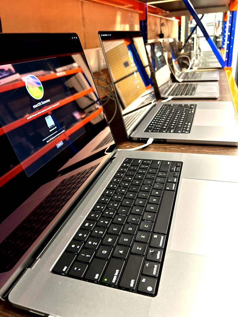 5 Silver MacBooks in a row on a wooden shelf with screens displaying a full factory reset.